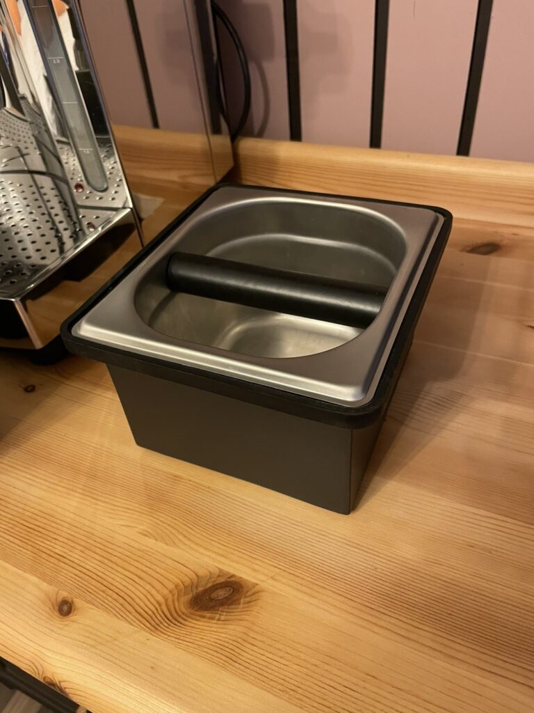 Eine hochwertige Knockbox aus Edelstahl neben einer Espressomaschine.